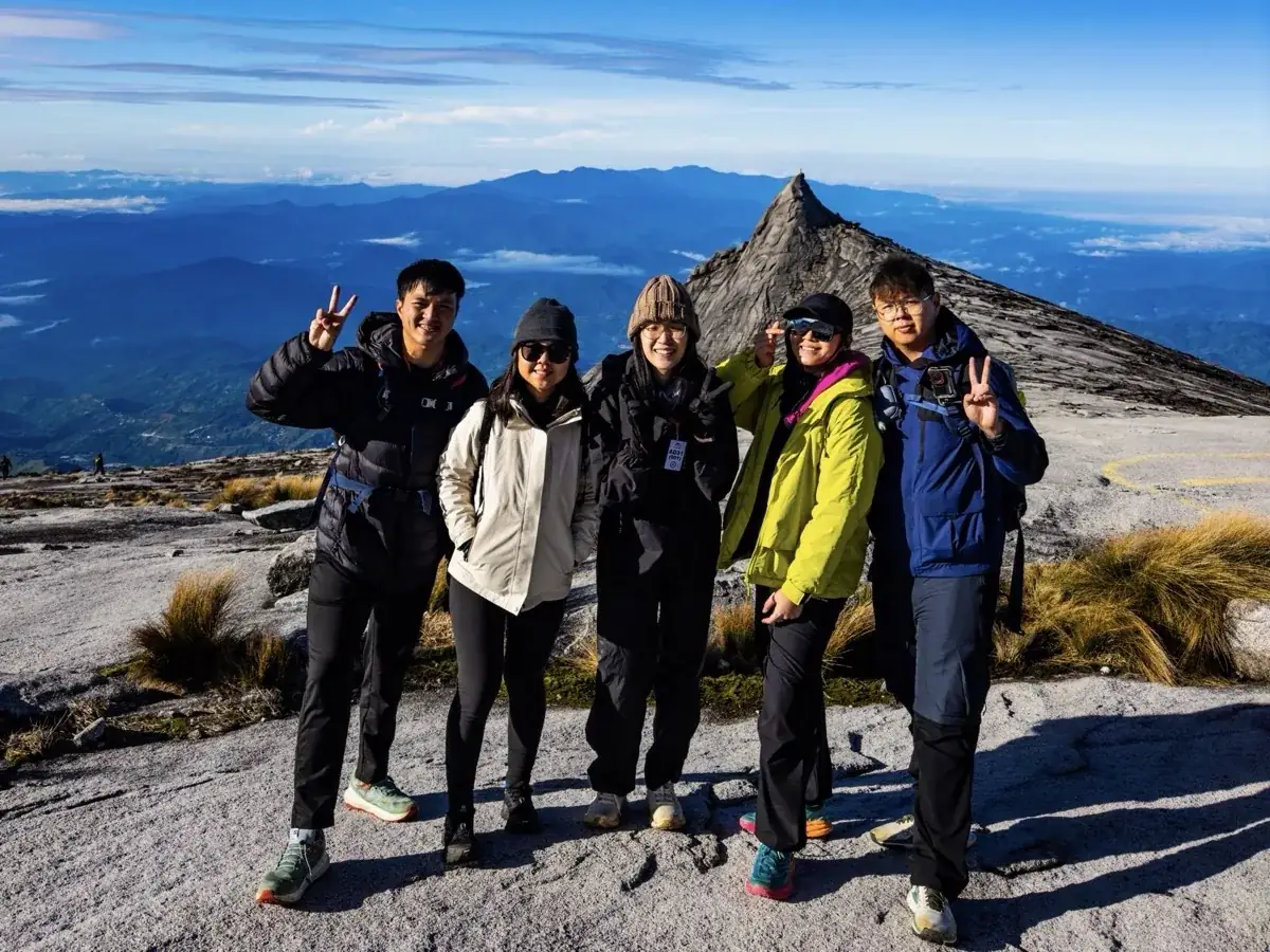 Five smiling climbers in warm jackets making peace signs on the summit plateau, South Peak behind