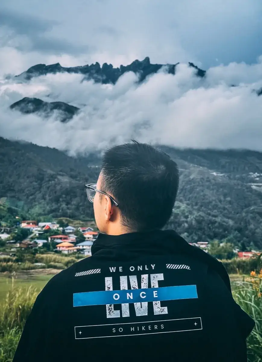 A So Hikers climber in a WE ONLY LIVE ONCE hoodie looking over Kundasang's houses to Kinabalu's jagged peaks above the clouds