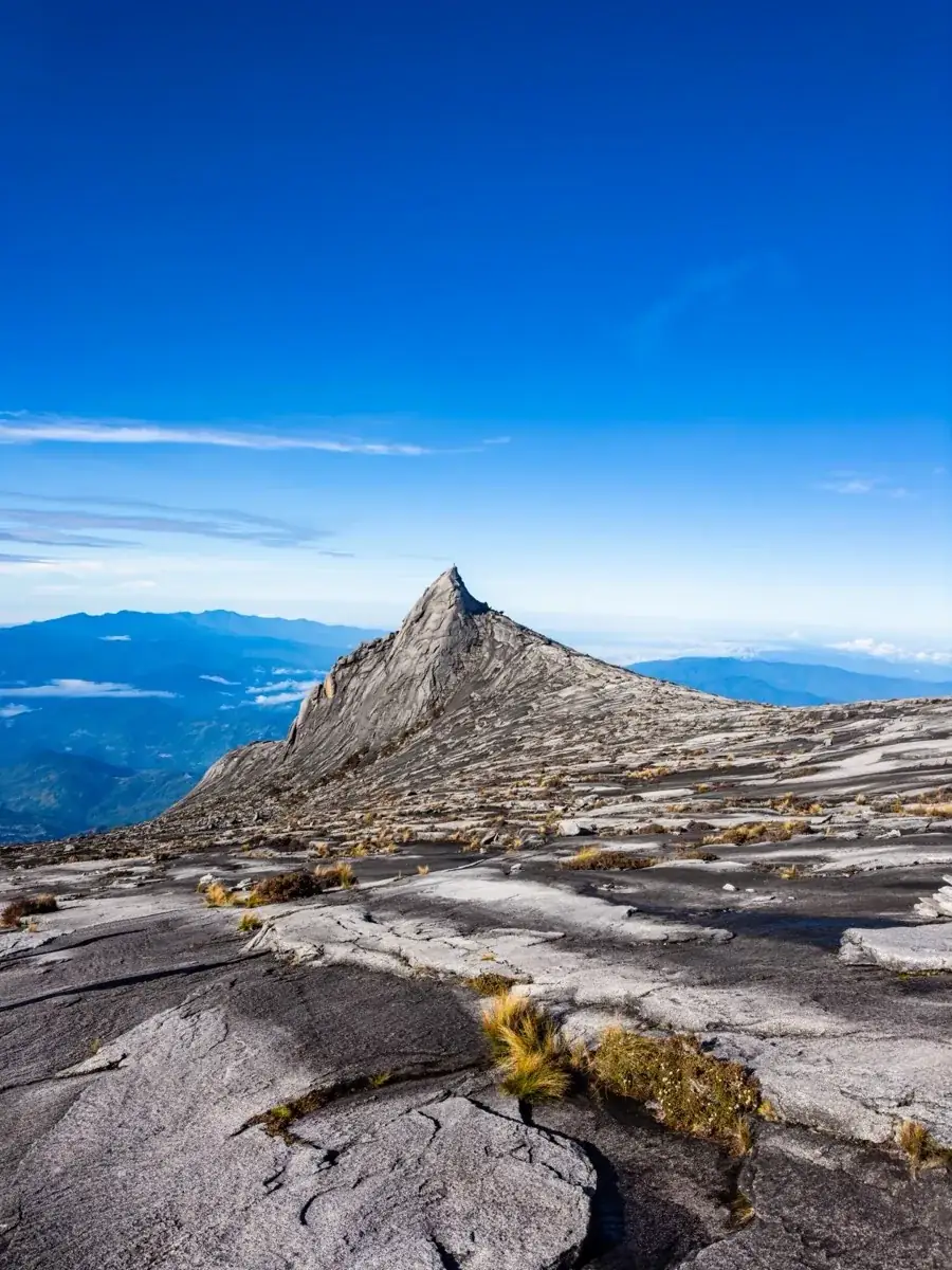South Peak's granite pyramid rising from the summit plateau under a blue sky, a sea of cloud below