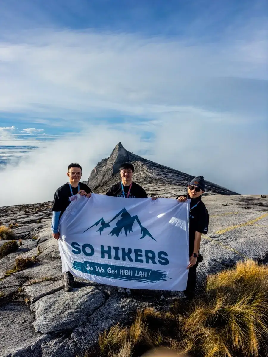 Three So Hikers climbers holding a SO HIKERS banner on the granite summit plateau of Mount Kinabalu, South Peak behind them above the clouds