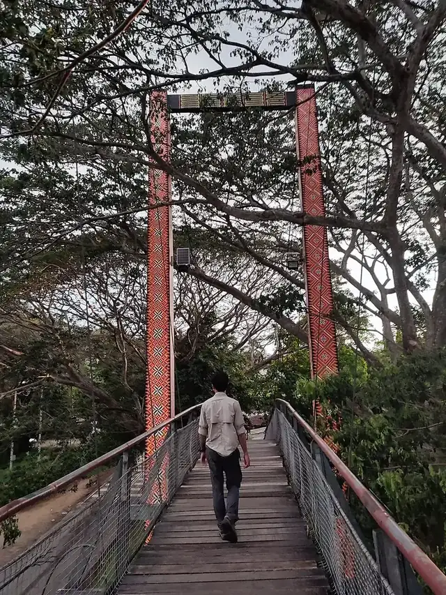 A man walking across the red-framed wooden Tamparuli suspension bridge under tall trees